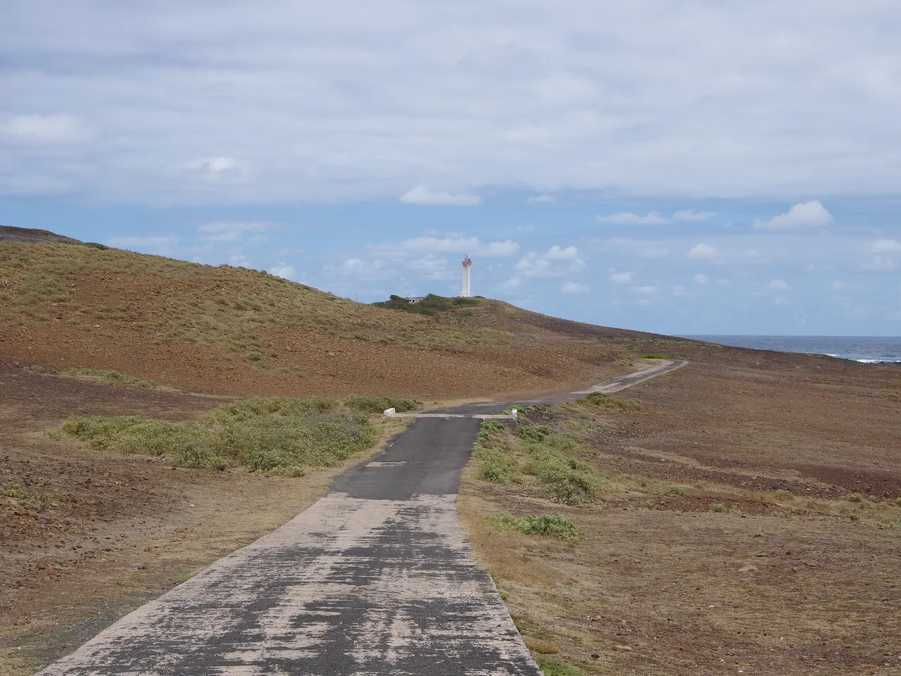 Une route étroite traverse le plateau aride et brun de La Désirade vers un phare blanc et rouge, avec l’océan à l’horizon.