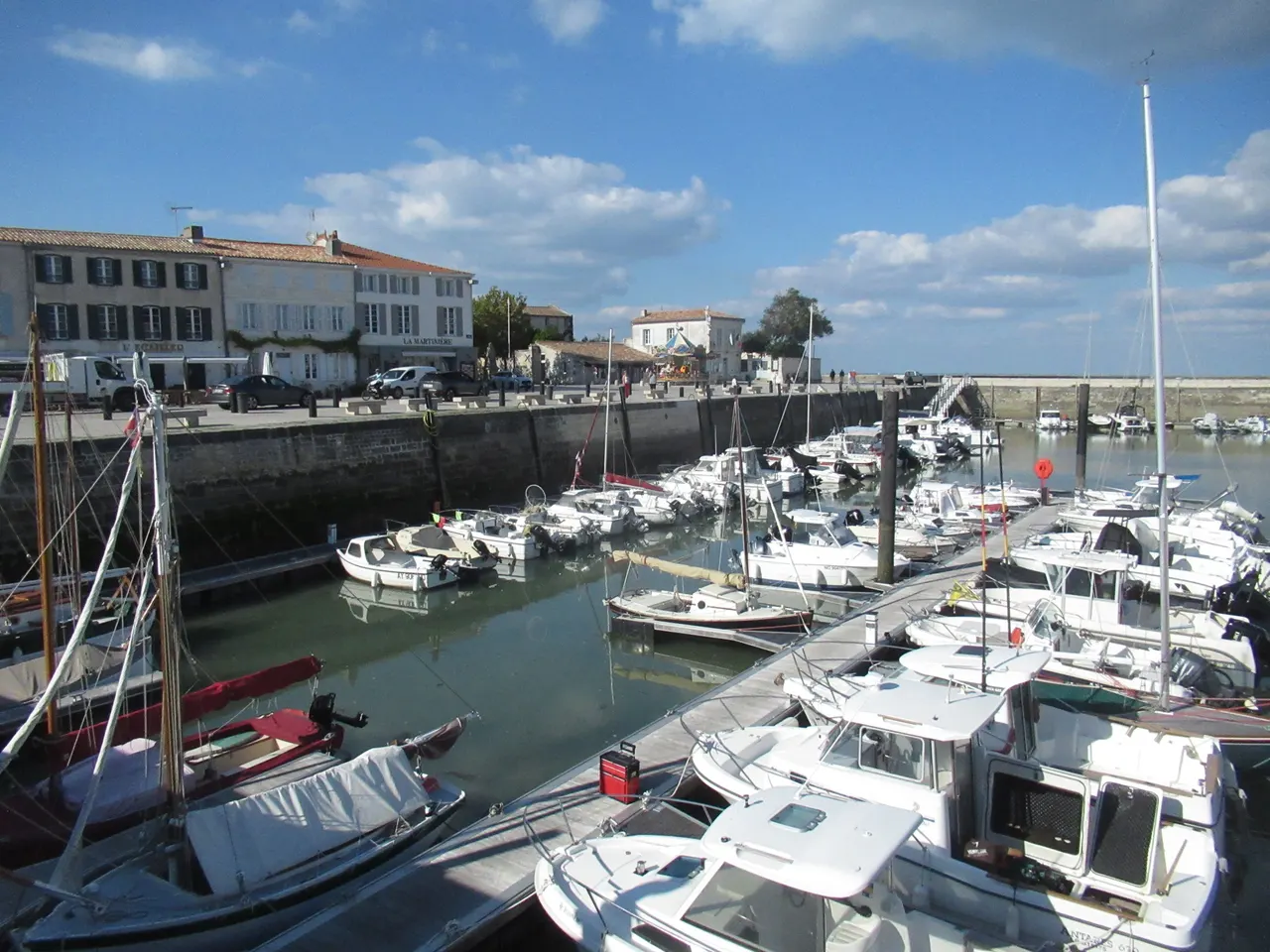 Le port de plaisance de La Flotte, ses pontons chargés de bateaux et son quai bordé de maisons blanches sous un ciel bleu nuageux.