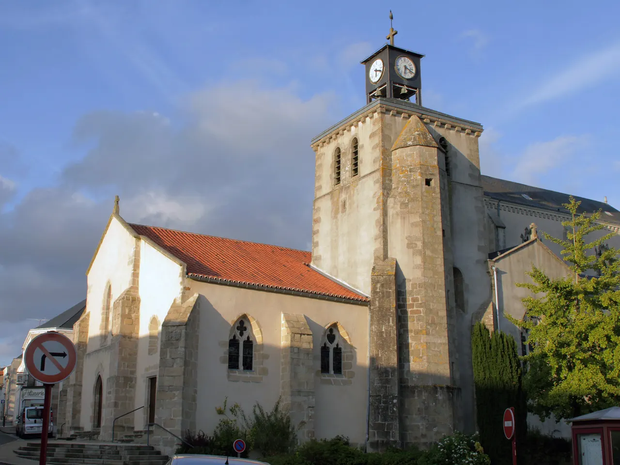 L’église Notre-Dame de La Séguinière, nef au toit de tuiles et tour de pierre surmontée d’un campanile à horloge, dans la lumière du soir.
