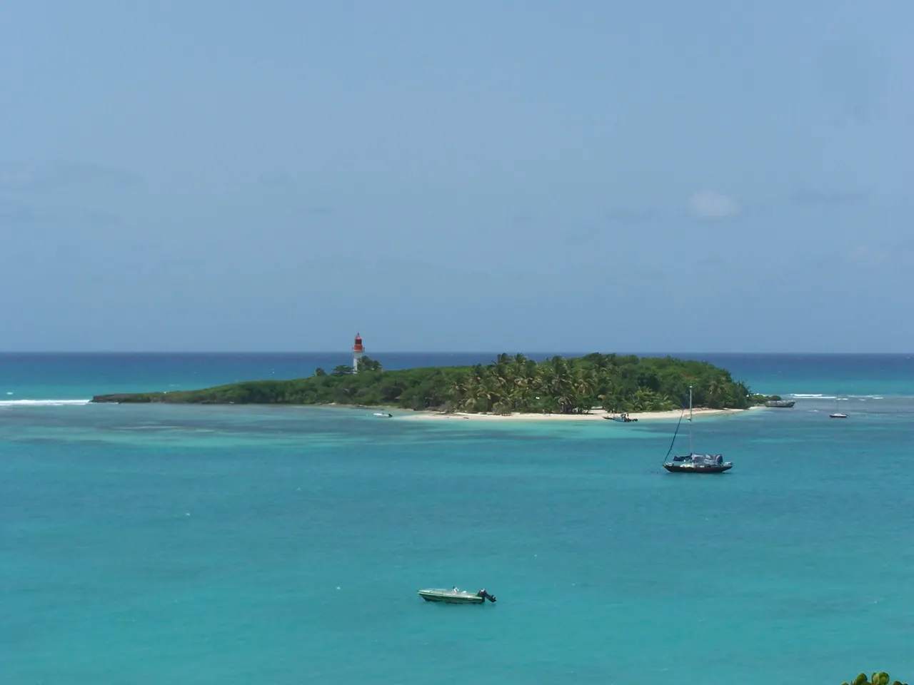L’îlet du Gosier et son phare rouge et blanc au milieu d’une mer turquoise, avec un voilier et une barque au mouillage.