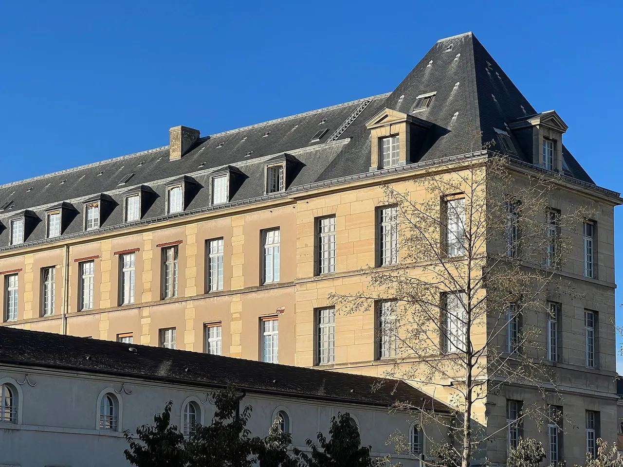 Un bâtiment classique de l’hôpital Bicêtre : façade de pierre blonde, haut toit d’ardoise percé de lucarnes et arbre aux feuilles clairsemées, sous un ciel bleu.
