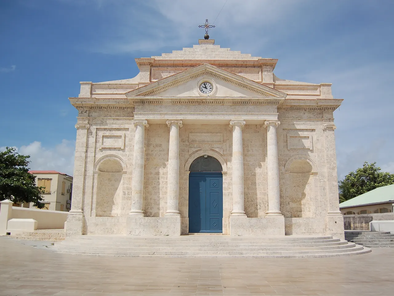La façade néoclassique en pierre claire de l’église Saint-Jean-Baptiste du Moule, avec ses quatre colonnes, son fronton à horloge et sa porte bleue.