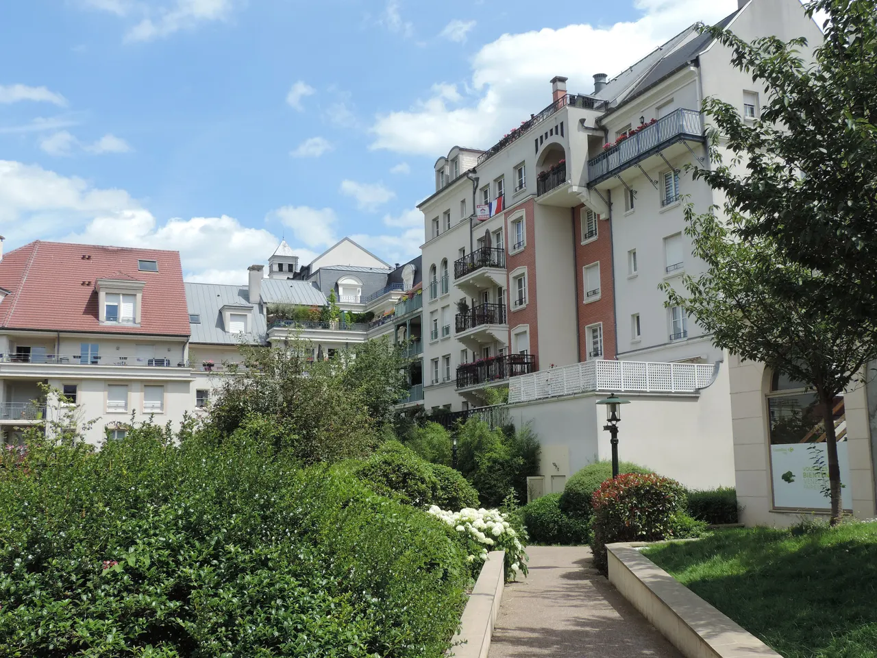 Immeubles clairs aux balcons fleuris du Cœur de Ville du Plessis-Robinson, au-dessus d’un jardin planté d’arbustes.