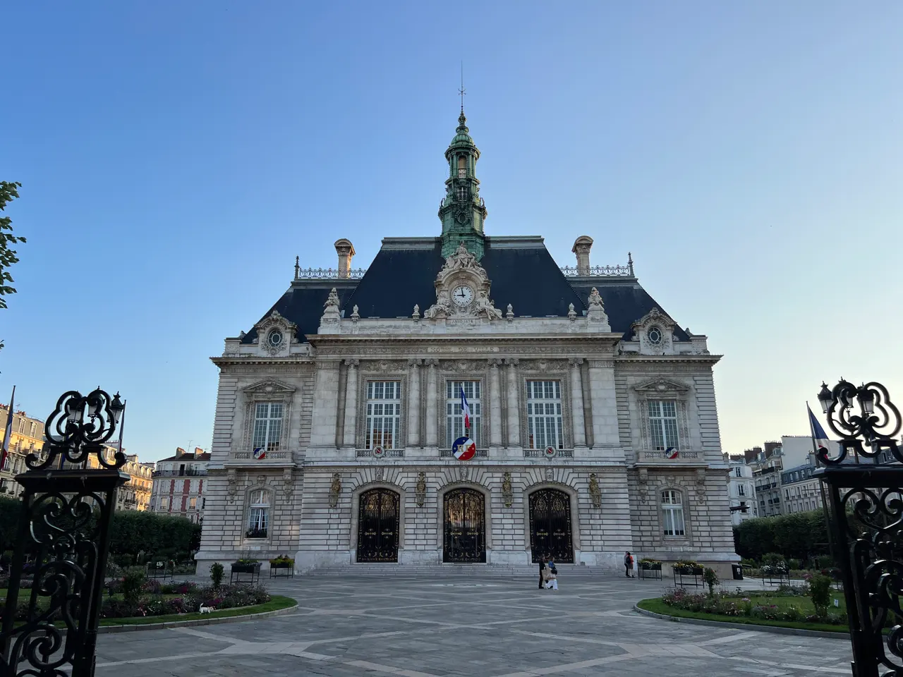L’hôtel de ville de Levallois-Perret, façade de pierre blanche et campanile vert-de-gris, vu entre les grilles de son square en fin de journée.