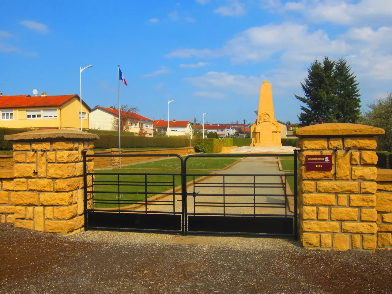 Entrée de la nécropole nationale de Lexy : piliers de pierre ocre, grille noire et allée menant à l’ossuaire, drapeau français à gauche.