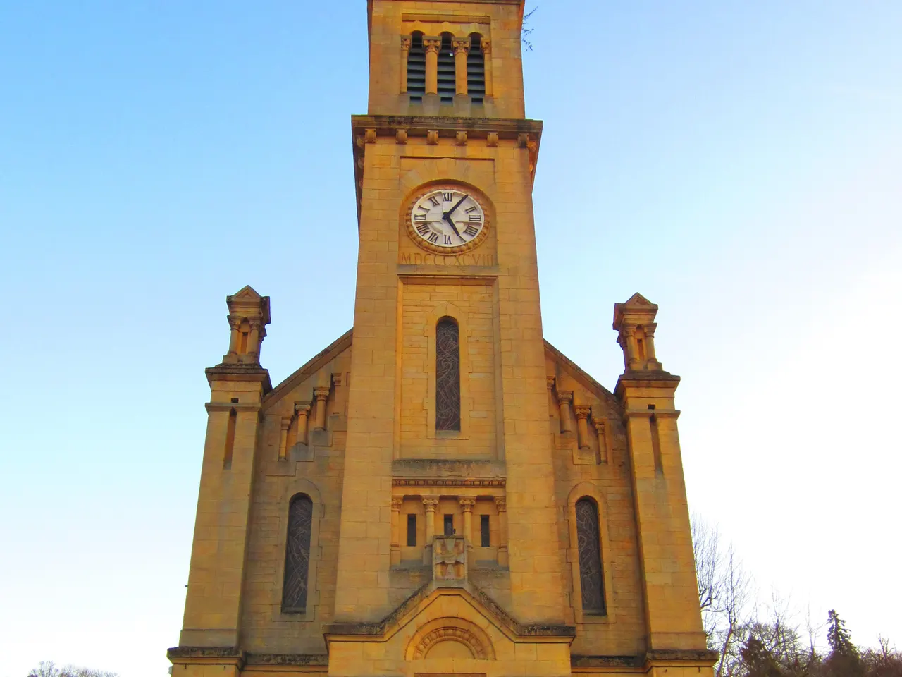 Clocher en pierre jaune de l’église Saint-Laurent de Longlaville, avec son horloge et la date MDCCCXCVIII gravée, sous un ciel bleu.