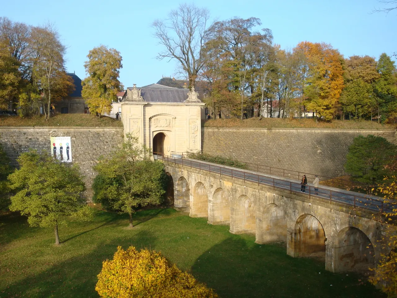 La porte de France de Longwy, au sommet des remparts de Vauban, reliée par un pont de pierre à arcades au-dessus d’un fossé herbeux, en automne
