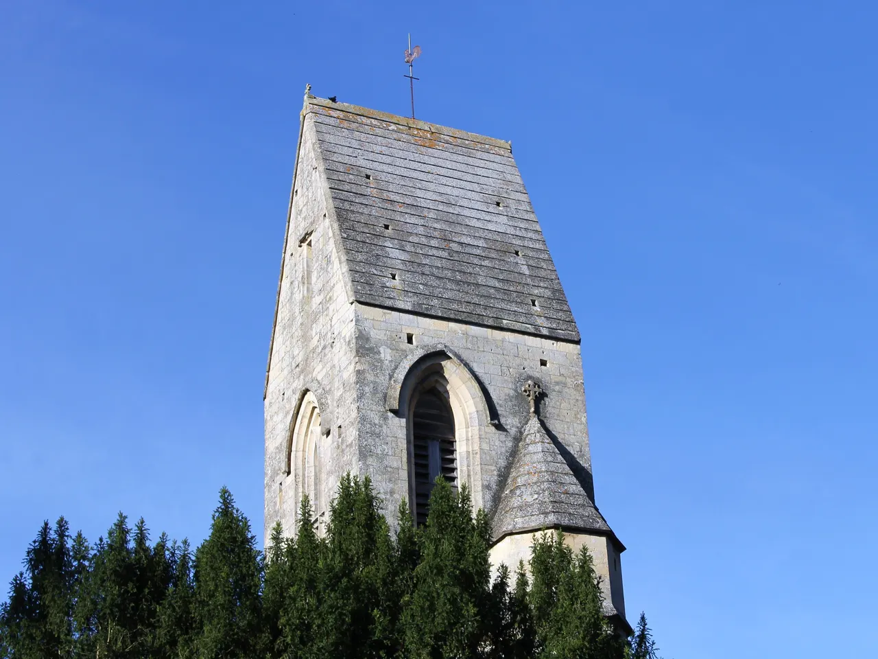 Le clocher en pierre de l’église Saint-Vigor de Louvigny, coiffé d’un toit en bâtière, au-dessus d’une rangée de conifères sous un ciel bleu