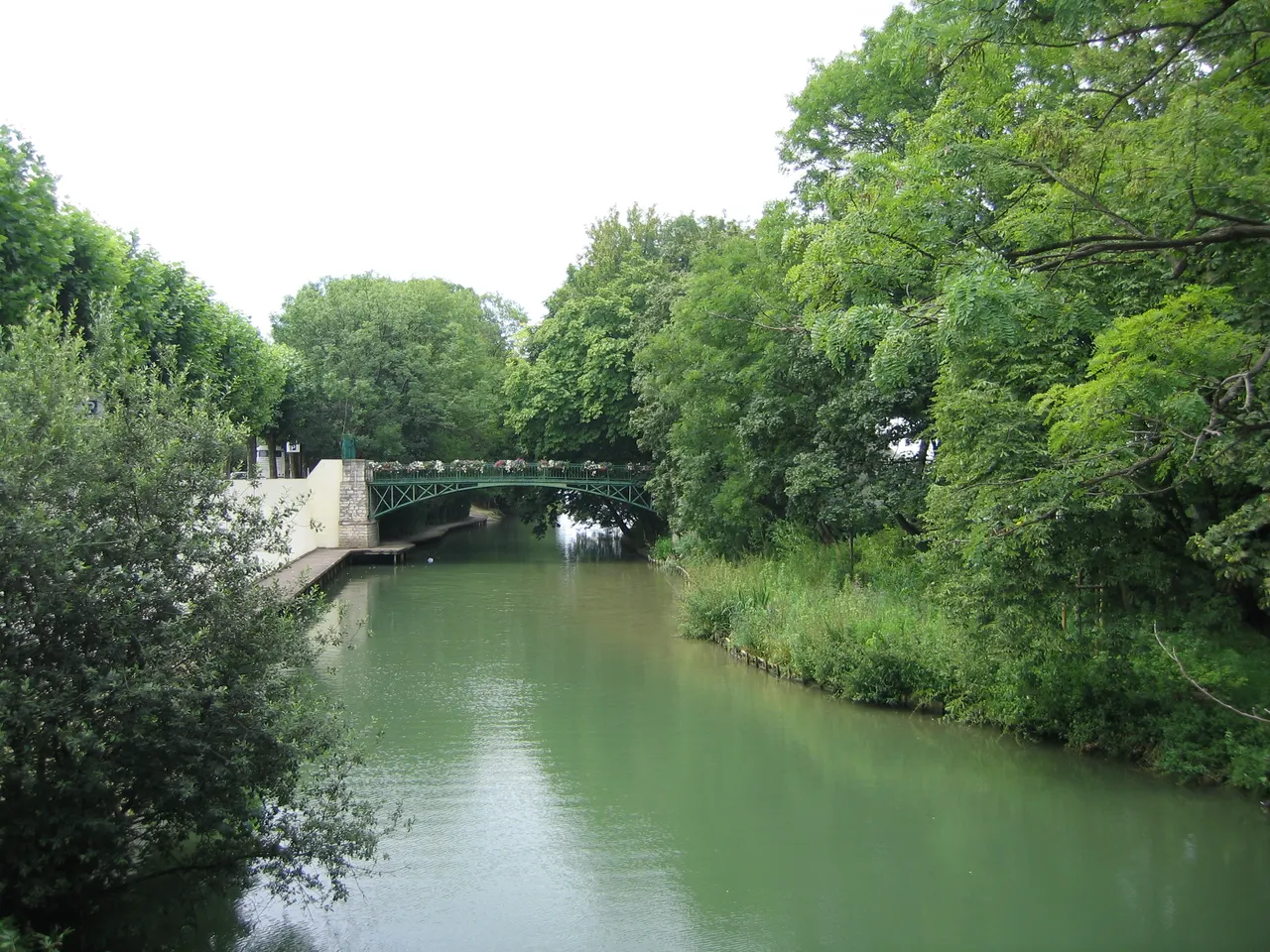 Un bras de la Marne à Maisons-Alfort, enjambé par une passerelle métallique verte garnie de fleurs, entre les arbres de la berge et l’île de Charentonneau à droite.