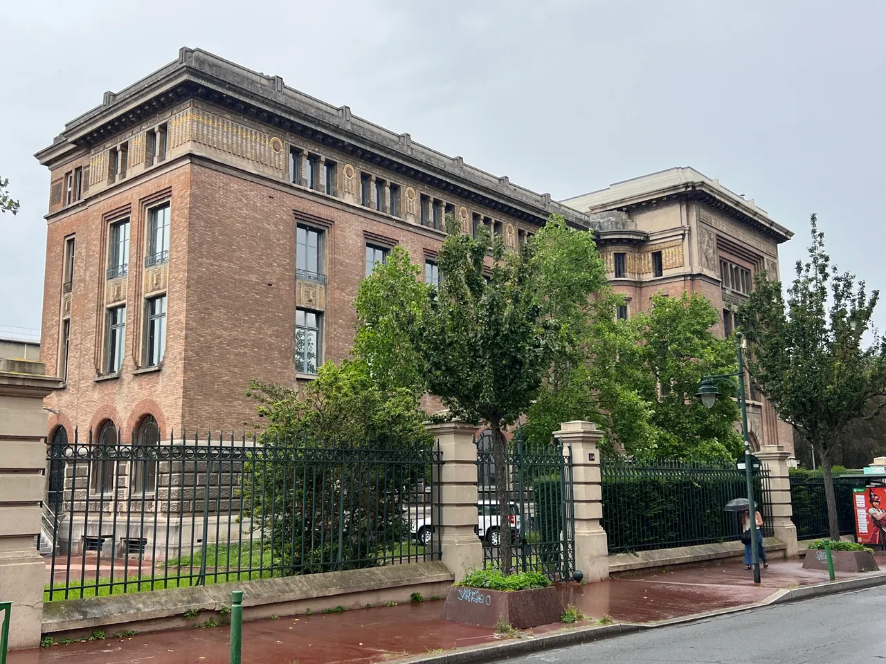 L’ancienne École supérieure d’électricité de Malakoff, grand bâtiment de brique aux frises ornées, derrière une grille et de jeunes arbres, sous un ciel gris.
