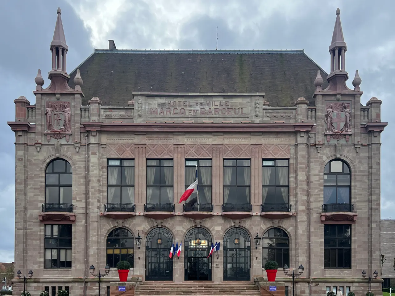 La façade de pierre rose de l’hôtel de ville de Marcq-en-Barœul, avec ses deux clochetons, ses grandes baies et ses drapeaux
