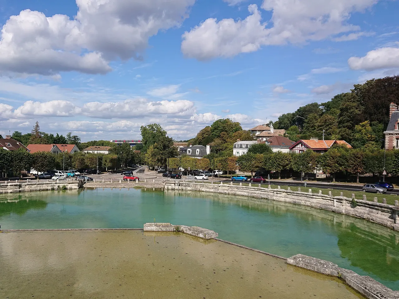 Le grand bassin de l’abreuvoir de Marly-le-Roi, son eau verte et ses bordures de pierre, avec des maisons et des arbres en arrière-plan sous un ciel nuageux.