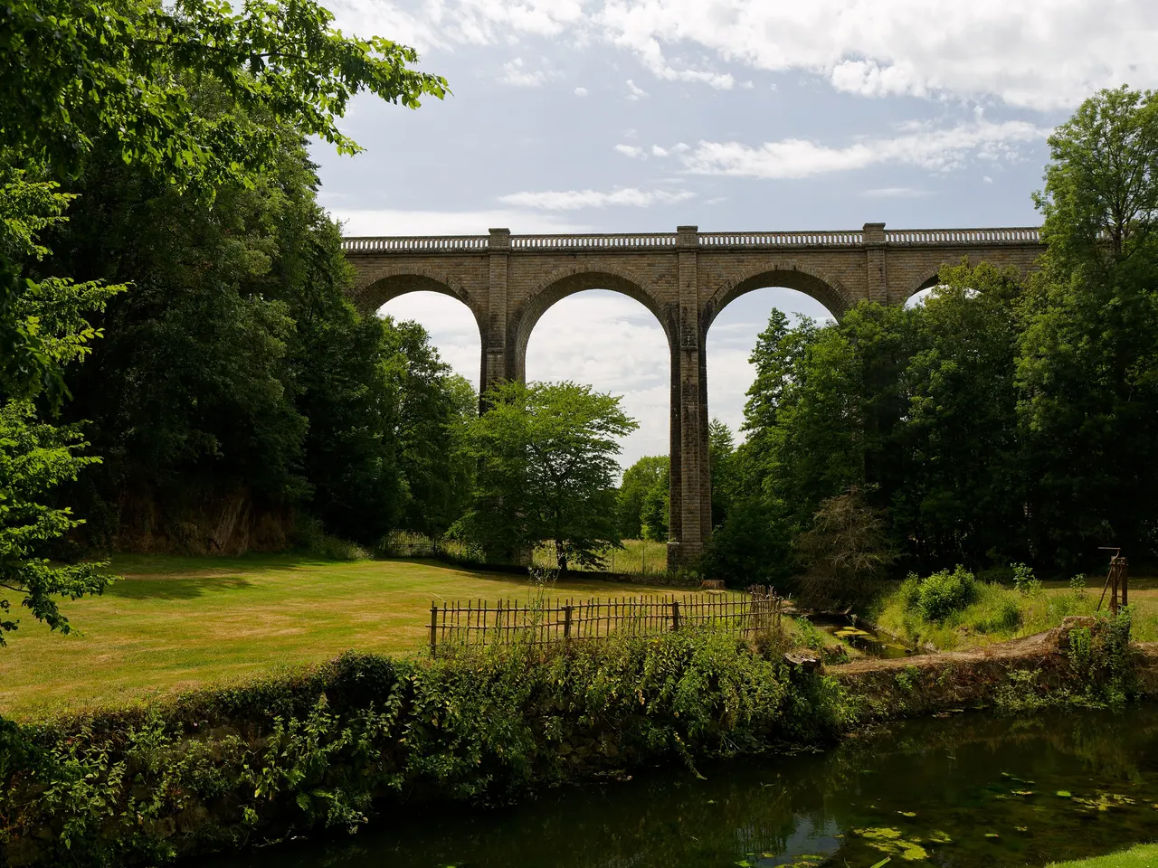 Un viaduc de pierre à grandes arches vu depuis une pelouse du parc oriental de Maulévrier, au bord de l’eau