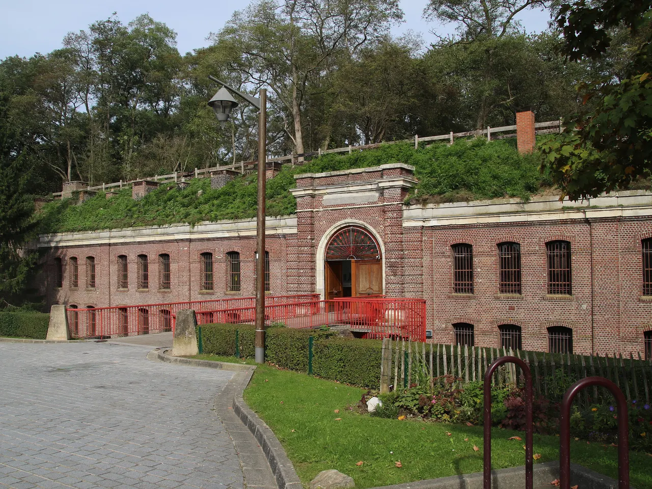 L’entrée du fort de Mons, façade de briques à fenêtres grillagées sous un talus herbeux, avec sa passerelle rouge menant au portail
