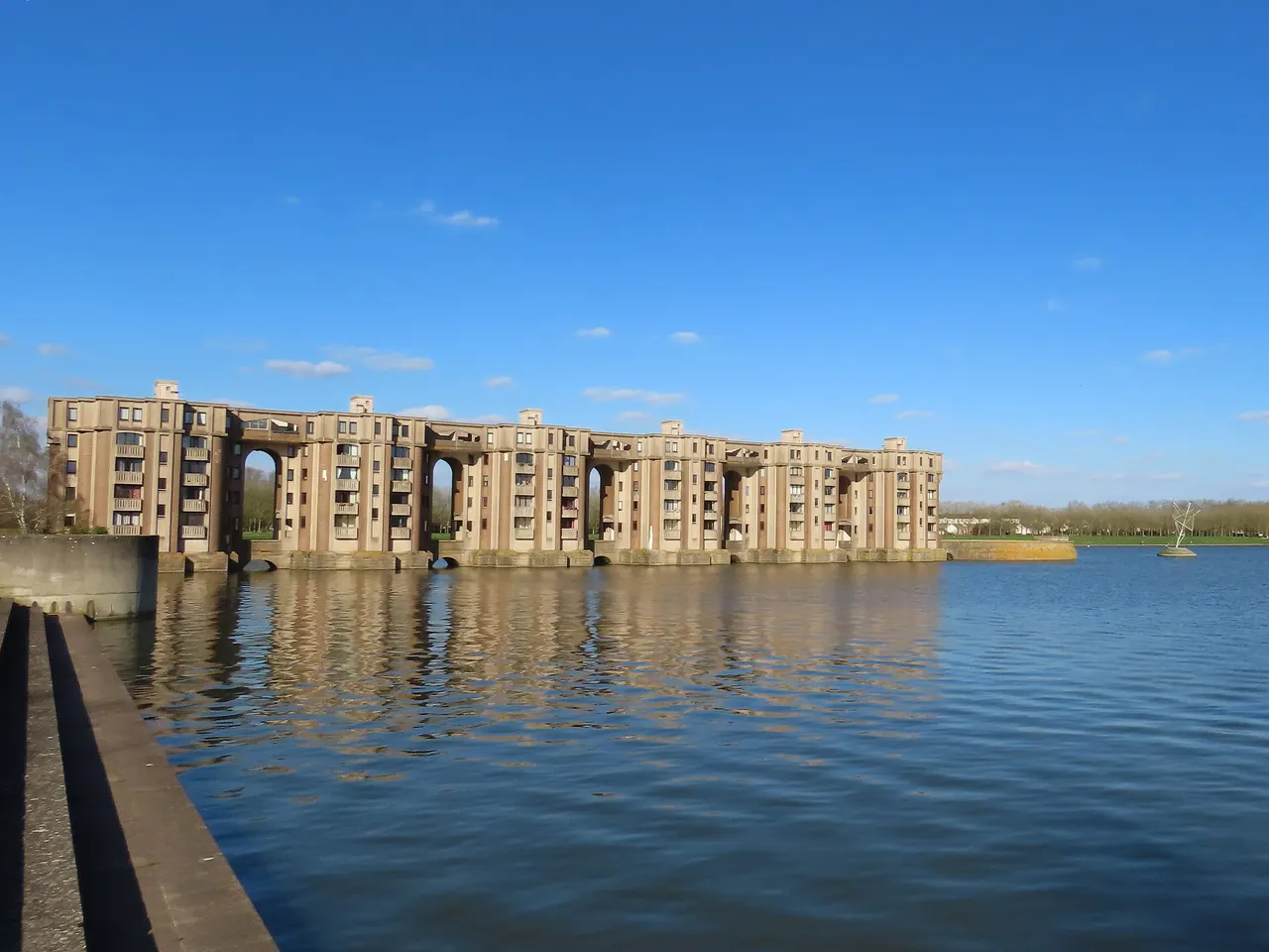 Les Arcades du Lac de Ricardo Bofill, longue façade percée de grandes arches, et leur reflet dans le bassin de la Sourderie sous un ciel bleu.