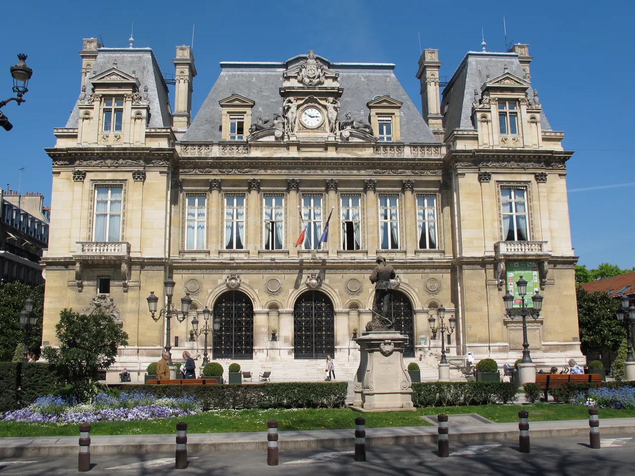 L’hôtel de ville de Neuilly-sur-Seine, façade de pierre ornée d’une horloge sous des toits d’ardoise, précédé d’un square fleuri et d’une statue sur son socle.
