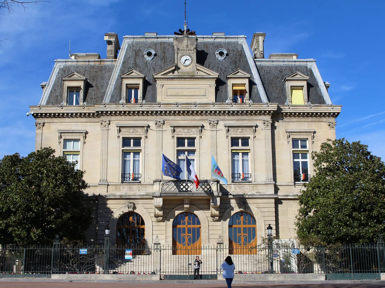 Mairie de Nogent-sur-Marne, façade en pierre à toit mansardé et horloge, encadrée de deux magnolias, sous un ciel bleu