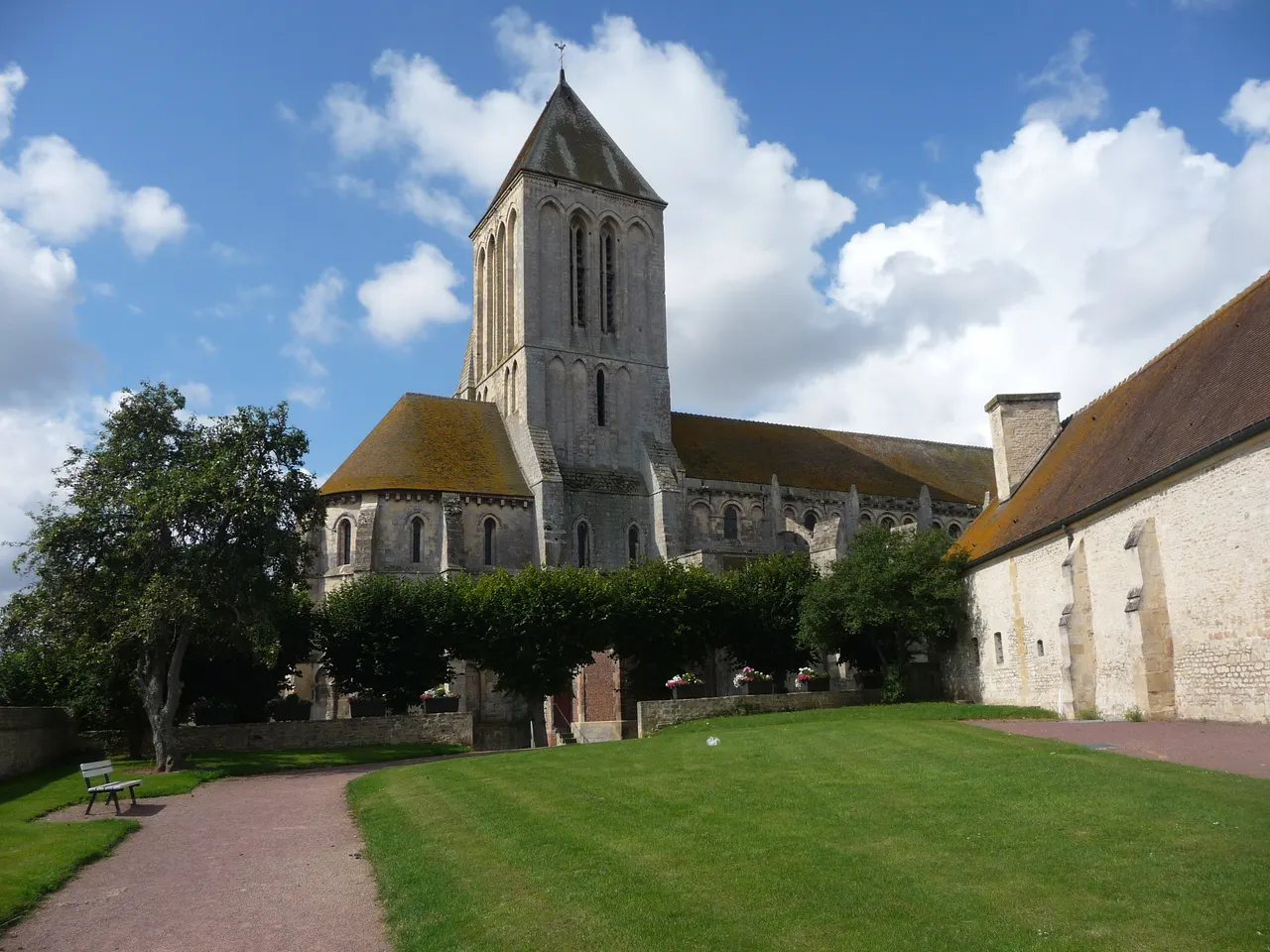 L’église Saint-Samson de Ouistreham, sa haute tour de pierre et son abside arrondie derrière un rideau d’arbres taillés, au bout d’une pelouse longée par un long bâtiment de pierre, sous un ciel bleu chargé de nuages blancs