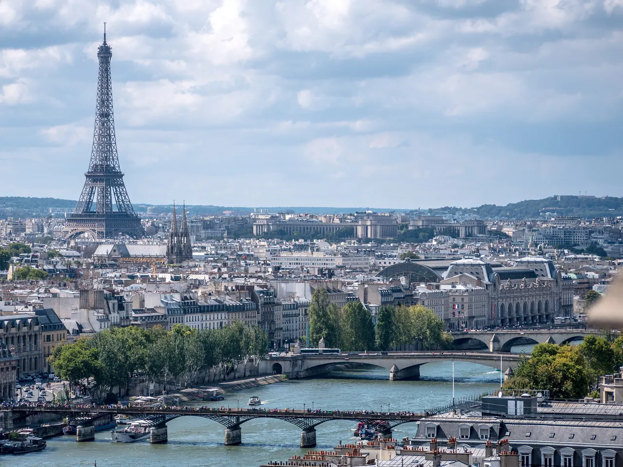 Paris vu d’en haut : la Seine et ses ponts au premier plan, le musée d’Orsay à droite, la tour Eiffel à gauche et les toits de la rive gauche sous un ciel nuageux.