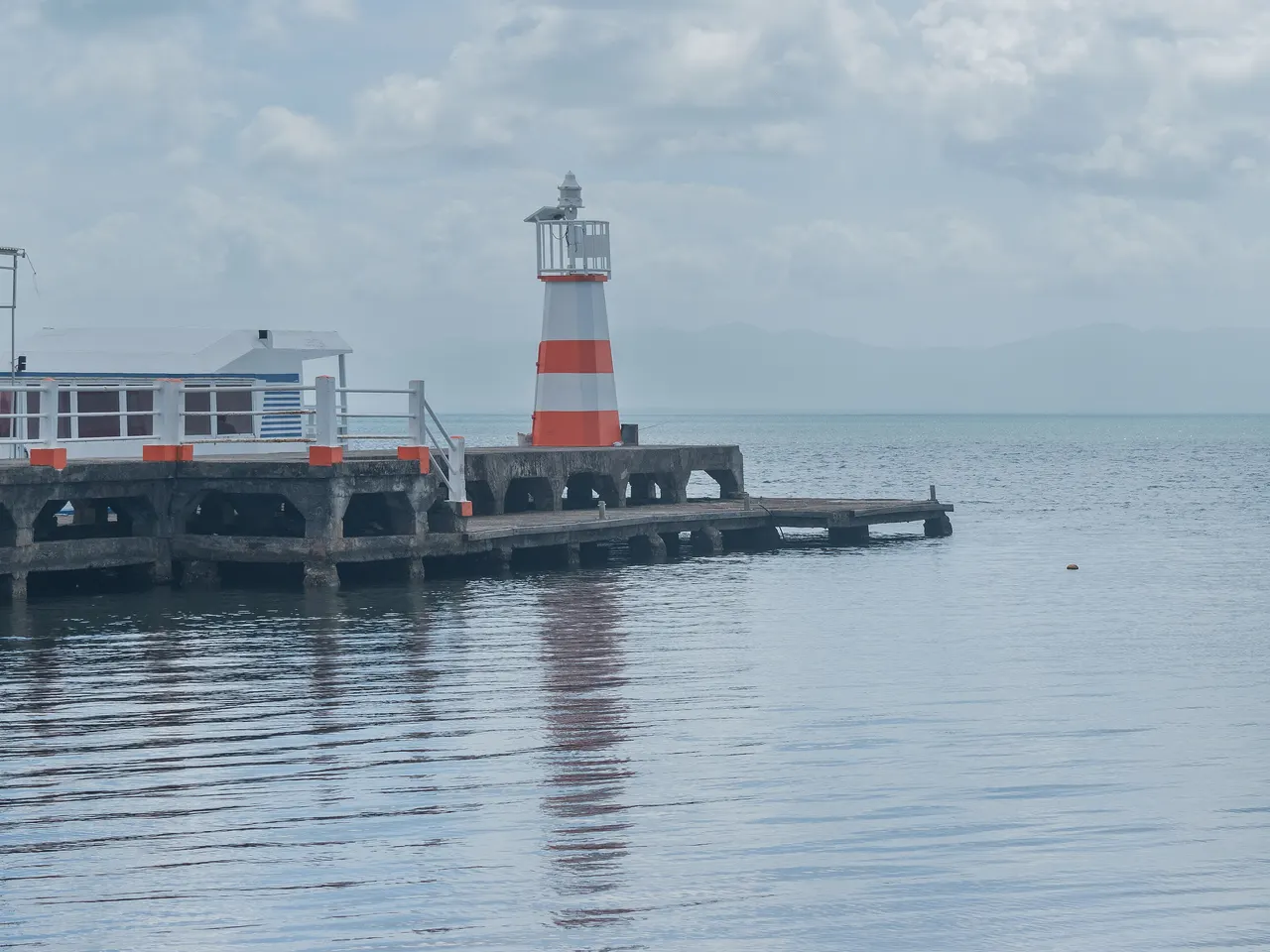 Le feu rayé de blanc et d’orange au bout du quai de béton de Petit-Canal, au-dessus des eaux calmes du Grand Cul-de-sac marin, avec des reliefs estompés par la brume à l’horizon