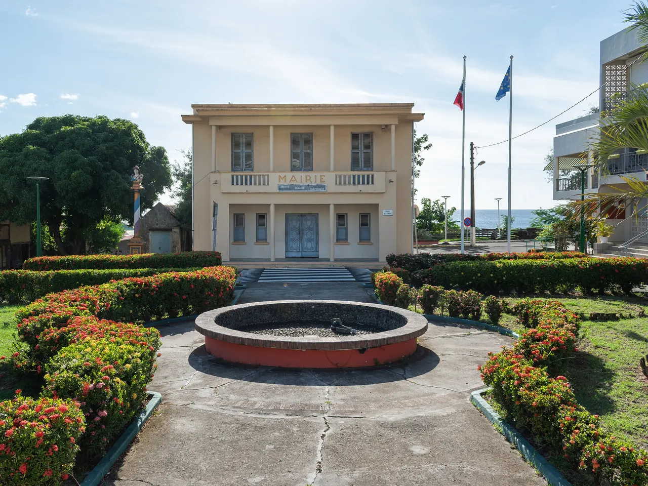 La mairie de Pointe-Noire dessinée par Ali Tur, derrière un bassin rond entouré de haies fleuries rouges ; à gauche la colonne républicaine surmontée d’un buste, au fond la mer.