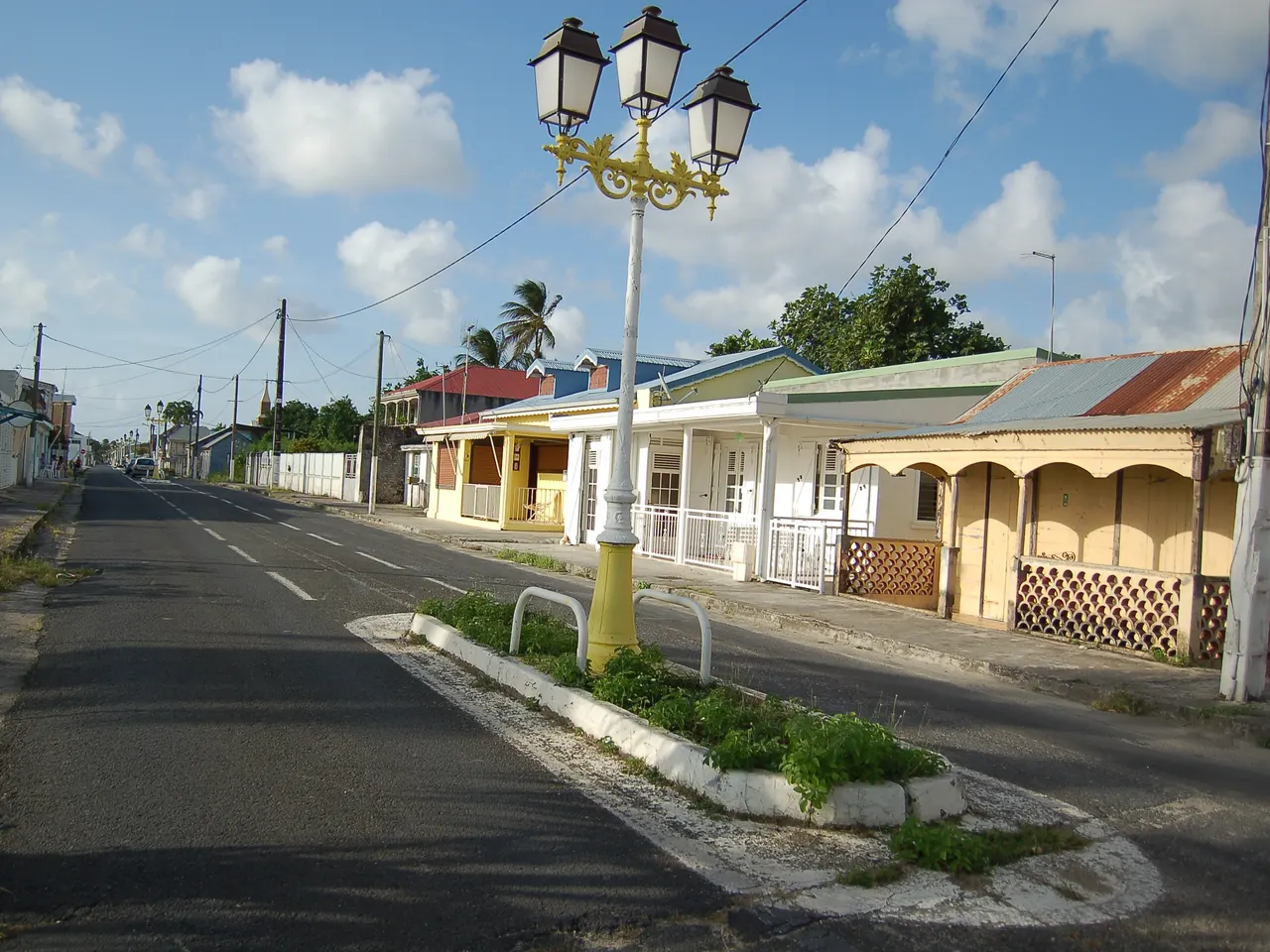 Une rue de Port-Louis bordée de maisons basses à galeries de bois et toits de tôle colorés, avec un lampadaire à trois lanternes, jaune et blanc, sur un terre-plein planté, sous un ciel bleu à nuages blancs