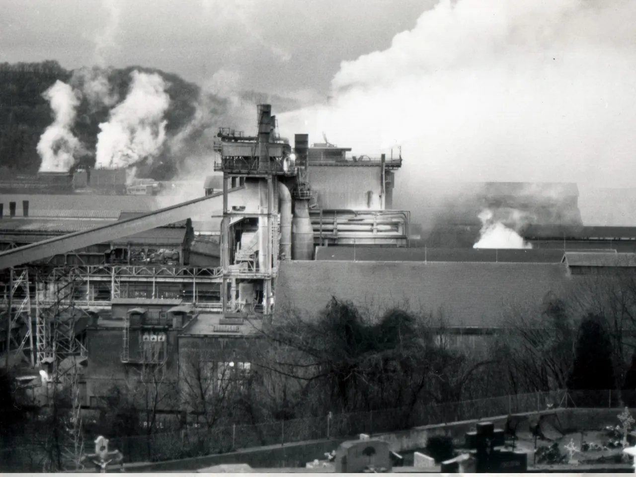 Photographie en noir et blanc de l’usine sidérurgique de la Providence à Réhon, ses installations noyées de fumée, avec des tombes au premier plan.