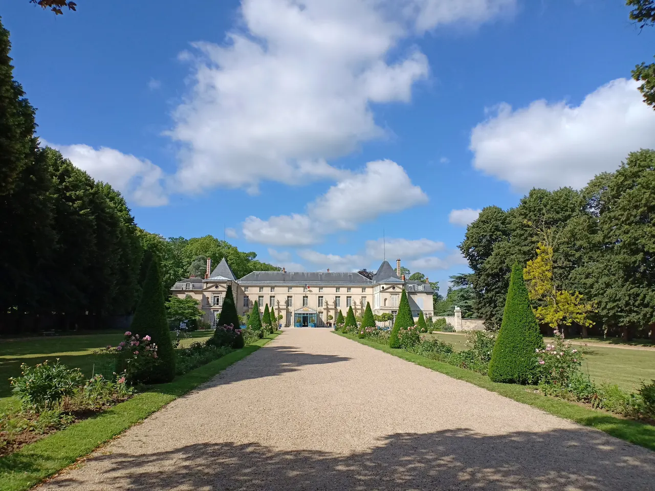 Le château de Malmaison au bout d’une allée bordée d’ifs taillés en cône et de rosiers, sous un ciel bleu.