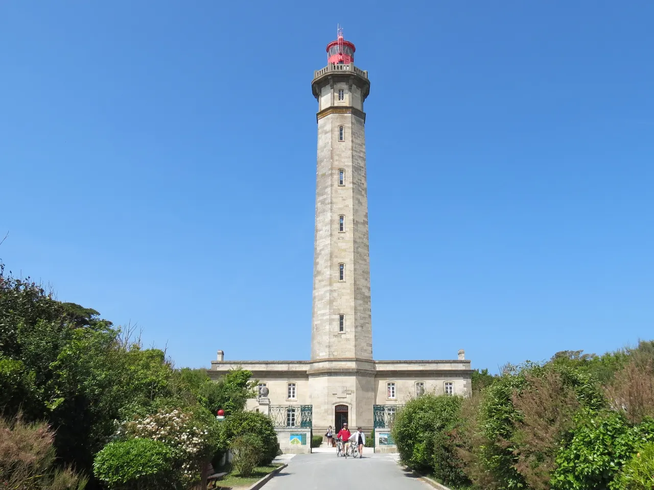 Le phare des Baleines à Saint-Clément-des-Baleines, haute tour de pierre à lanterne rouge dressée derrière son bâtiment d’accueil et des massifs verts.