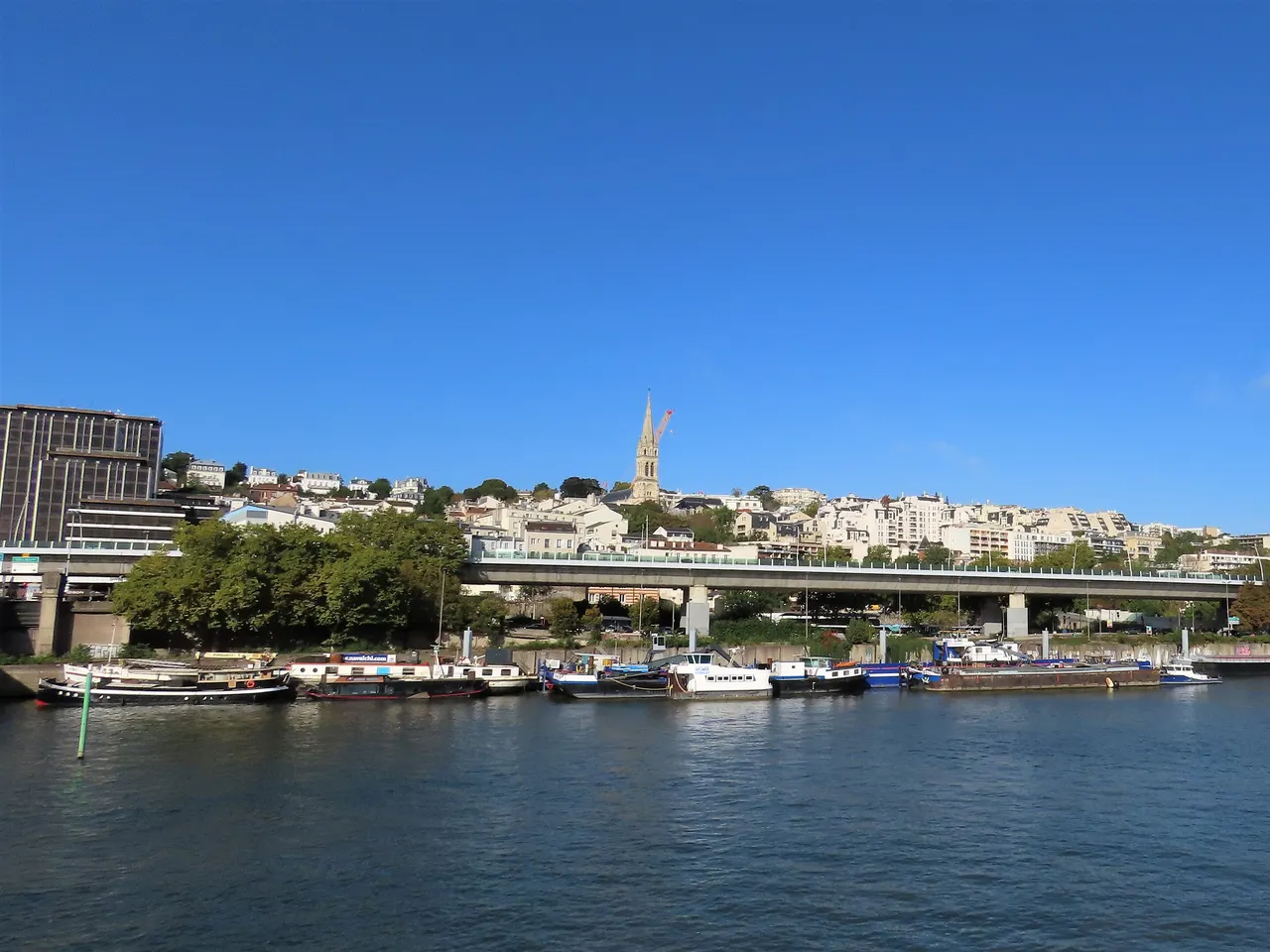 Saint-Cloud étagé sur son coteau au-dessus de la Seine, dominé par la flèche de l’église Saint-Clodoald, avec des péniches amarrées au quai.
