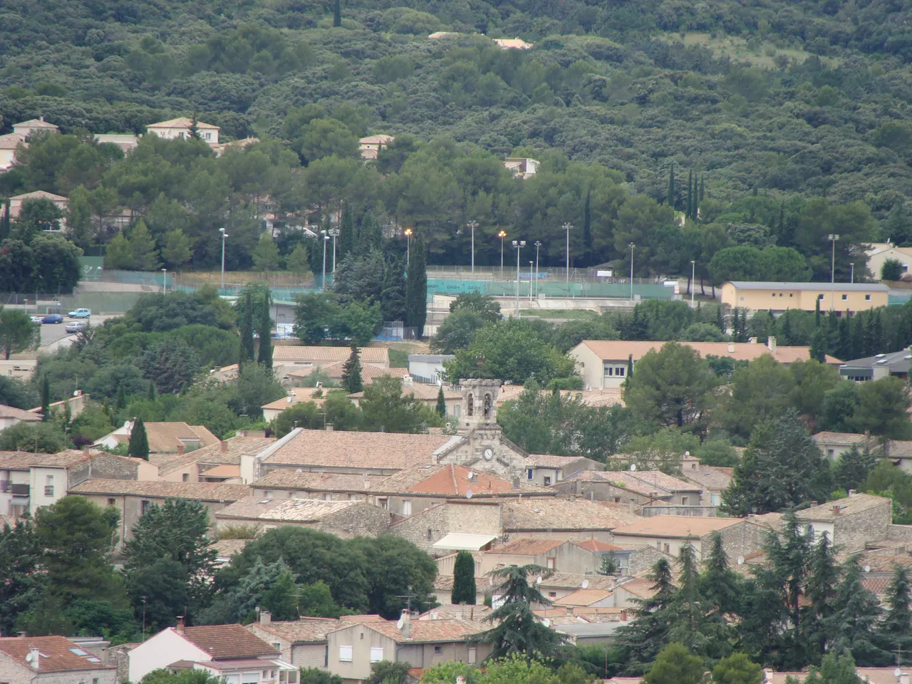 Les toits de tuiles du centre de Saint-Gély-du-Fesc autour d’un clocher à horloge, au pied d’une colline couverte de chênes et de pins.