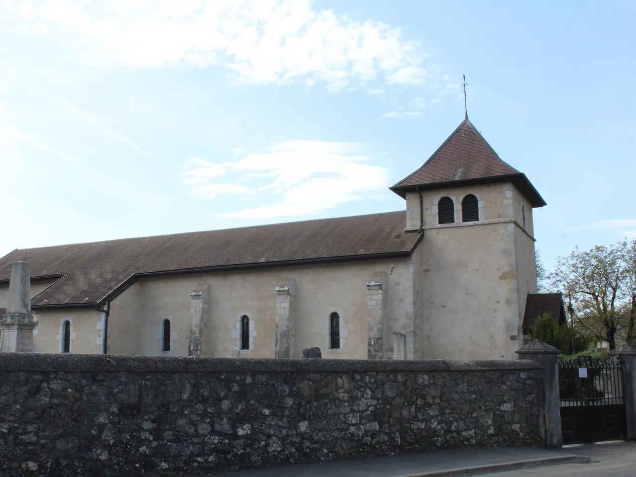 L’église Saint-Pierre de Pouilly, à Saint-Genis-Pouilly : nef enduite et clocher carré au toit de tuiles brunes, derrière un mur de pierre, sous un ciel bleu.