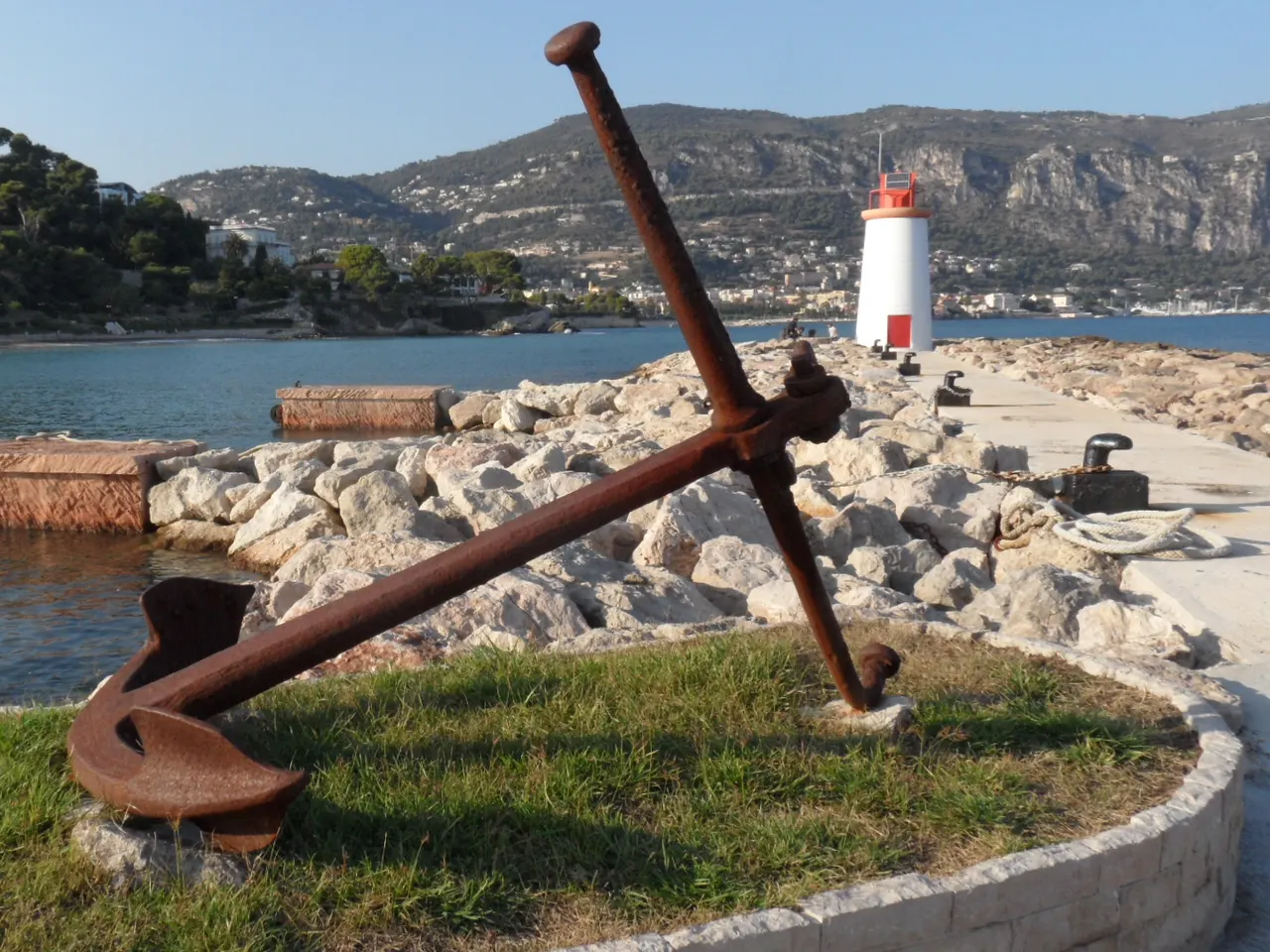 Une vieille ancre rouillée posée sur une pelouse au bord de la jetée du port de Saint-Jean-Cap-Ferrat, avec le petit phare blanc et rouge au bout de la digue et la côte montagneuse en arrière-plan.