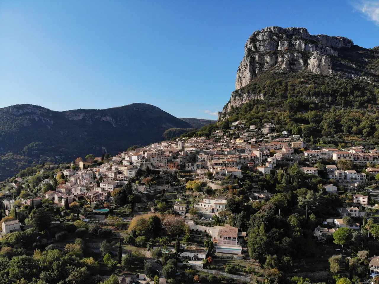 Saint-Jeannet étagé sur sa colline sous la haute falaise calcaire du Baou, dans un ciel bleu, avec d’autres reliefs boisés sur la gauche.