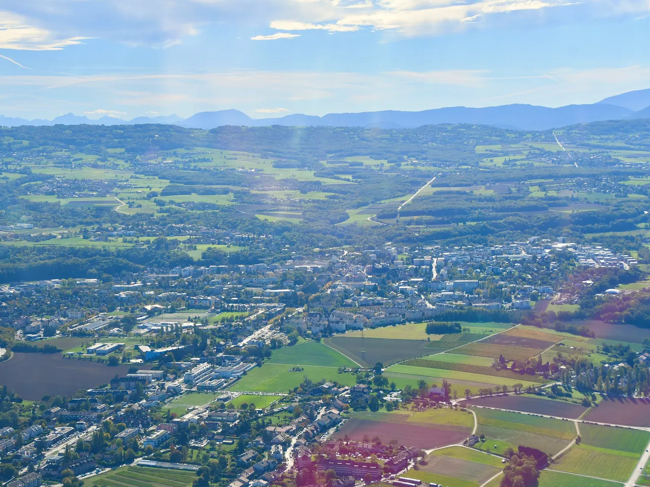 Vue aérienne de Saint-Julien-en-Genevois : immeubles et maisons entre des champs cultivés et des bois, collines puis montagnes bleutées à l’horizon.