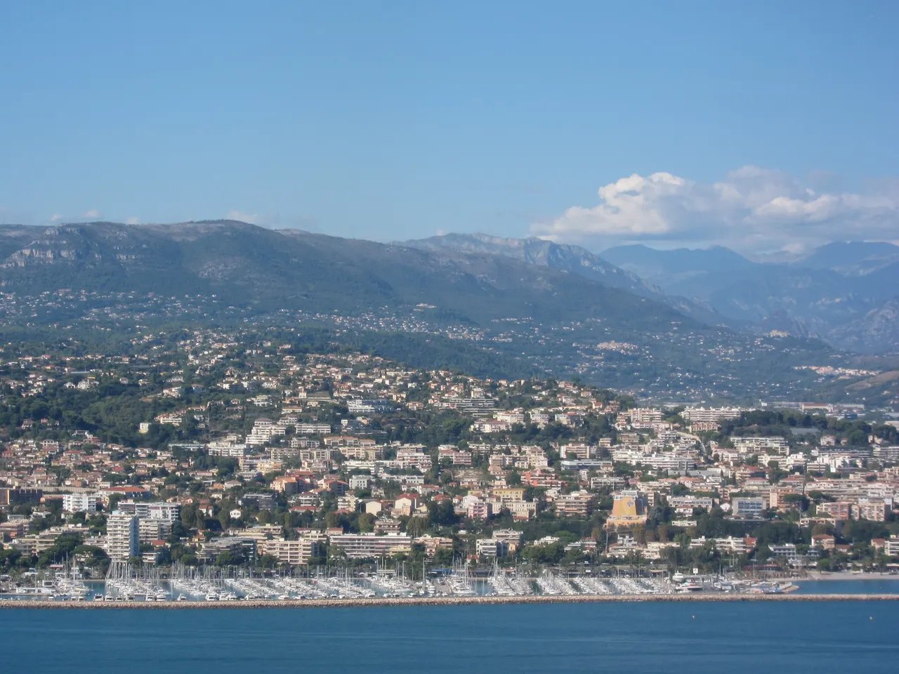 Saint-Laurent-du-Var vu d’avion : le port de plaisance derrière sa digue, la ville étagée sur les collines et les montagnes à l’arrière-plan.