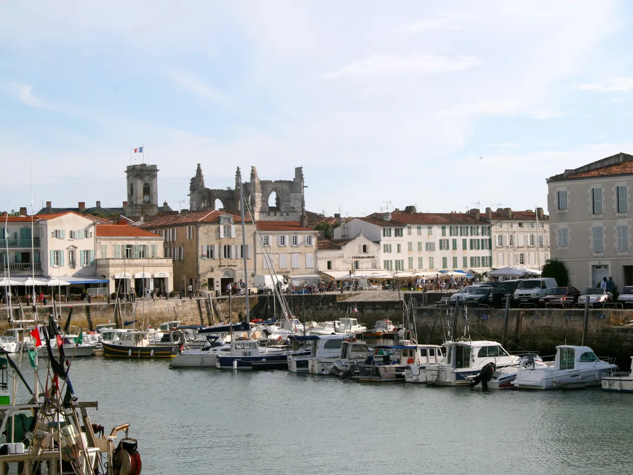 Le port de Saint-Martin-de-Ré, ses bateaux amarrés devant les façades claires des quais, avec le clocher et les ruines de l’église Saint-Martin au-dessus des toits.