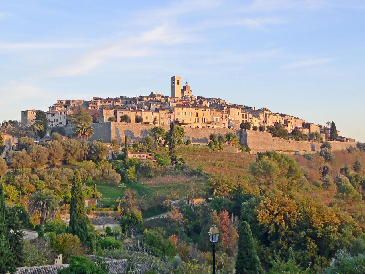 Le village perché de Saint-Paul-de-Vence dans une lumière dorée, ceint de ses remparts, au-dessus de terrasses plantées de cyprès et de palmiers.