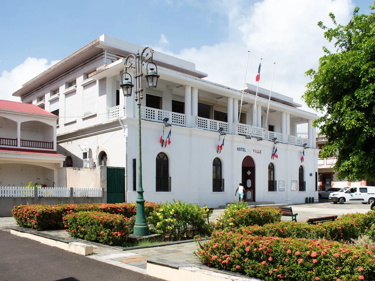 L’hôtel de ville de Sainte-Rose, façade blanche à fenêtres cintrées et galerie à balustres ajourés, pavoisée de drapeaux tricolores, derrière un réverbère vert et des massifs de fleurs rouges
