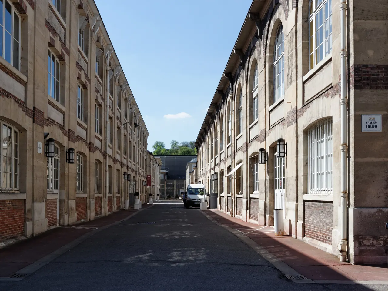Une allée de la Manufacture nationale de Sèvres entre deux longs bâtiments d’ateliers en pierre et brique, sous un ciel bleu.