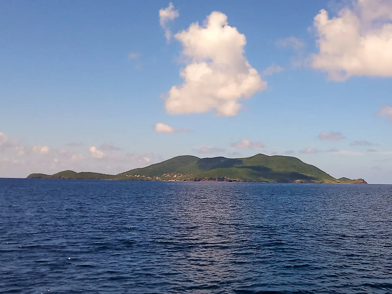L’île de Terre-de-Bas vue depuis la mer : des mornes verts piqués de quelques maisons, sous un ciel bleu traversé de nuages blancs