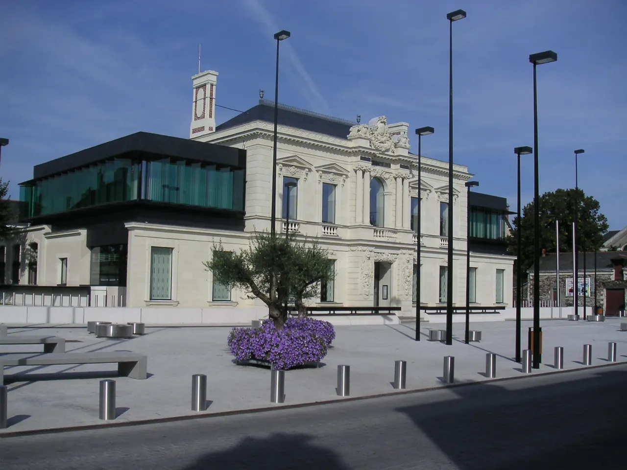 L’hôtel de ville de Trélazé, façade claire sculptée flanquée d’extensions vitrées, derrière une place dallée plantée d’un olivier et de fleurs violettes.