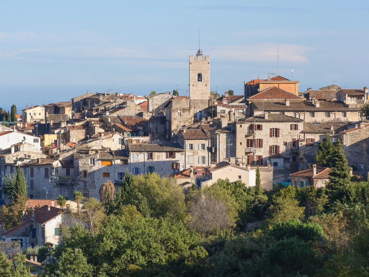Les maisons de pierre aux toits de tuiles de la vieille ville de Vence, dominées par un clocher carré, au-dessus d’une masse d’arbres.