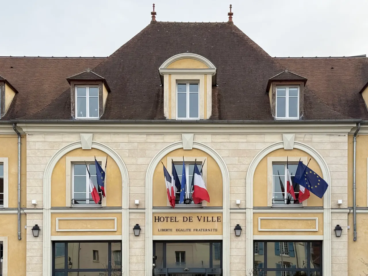 Façade jaune et pierre de l’hôtel de ville de Verrières-le-Buisson, trois arcades garnies de drapeaux sous un grand toit de tuiles brunes