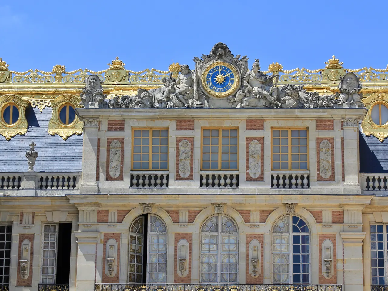 Façade de la cour de Marbre du château de Versailles, avec son horloge dorée encadrée de statues sous un ciel bleu
