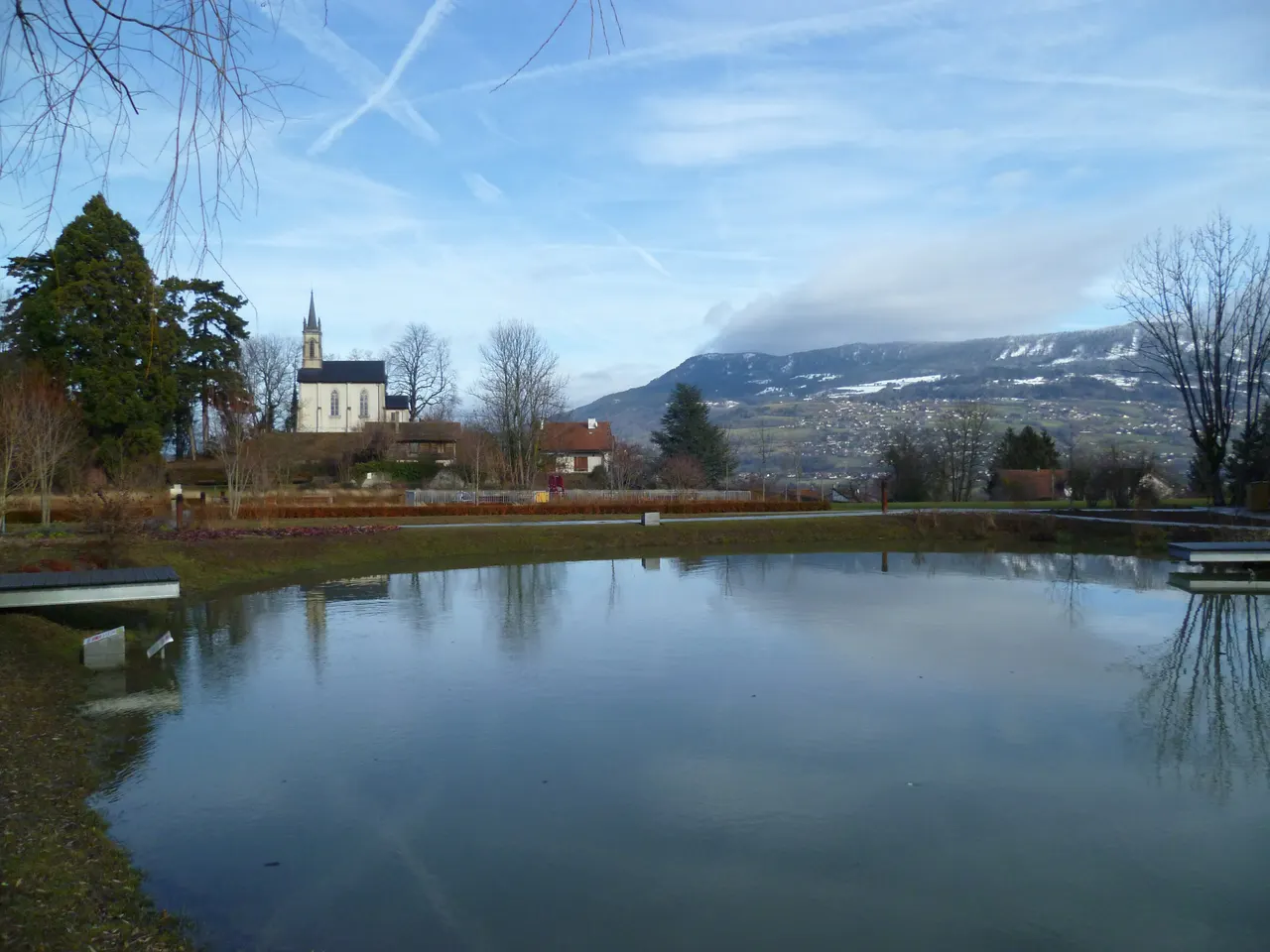 Un étang aux eaux calmes à Vétraz-Monthoux, une église néogothique sur une butte arborée et des pentes de montagne saupoudrées de neige à l’horizon.
