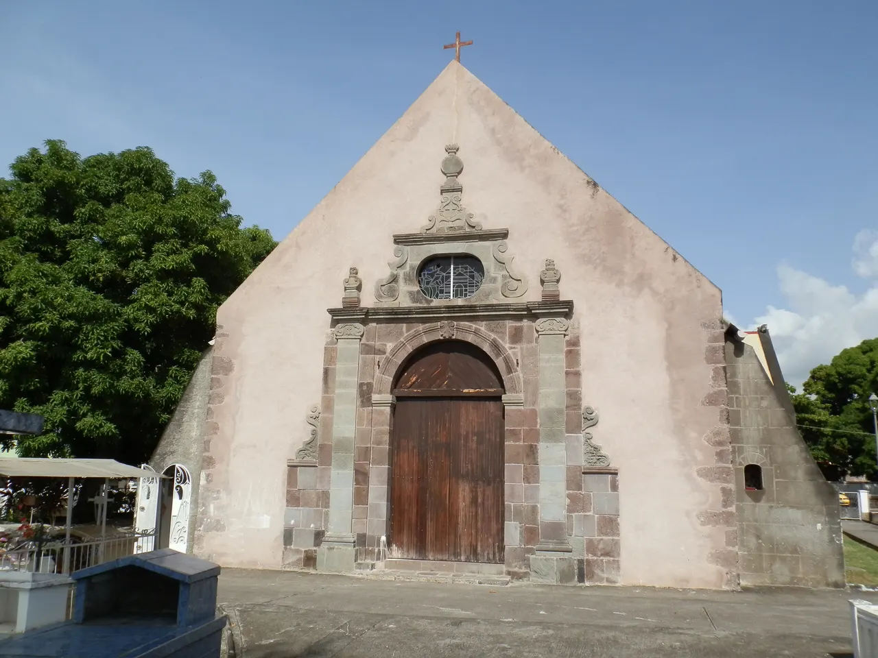 La façade de l’église Saint-Joseph de Vieux-Habitants : haut pignon enduit, portail en pierre de taille autour d’une porte de bois cintrée, oculus et croix au sommet ; tombes du cimetière au premier plan à gauche.
