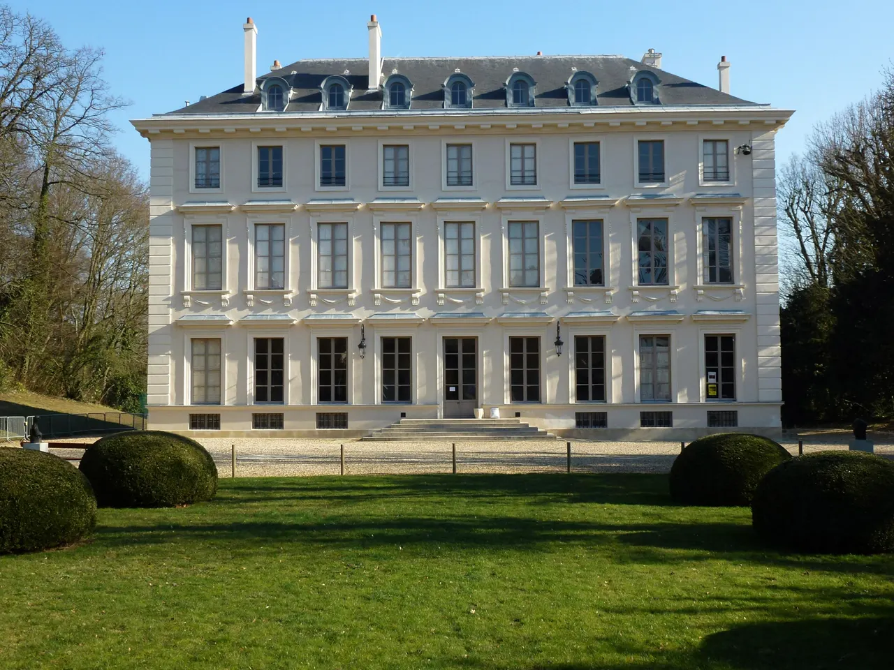 La façade claire du château de Thierry à Ville-d’Avray, devant une pelouse et des buis taillés en boule, entre de grands arbres.