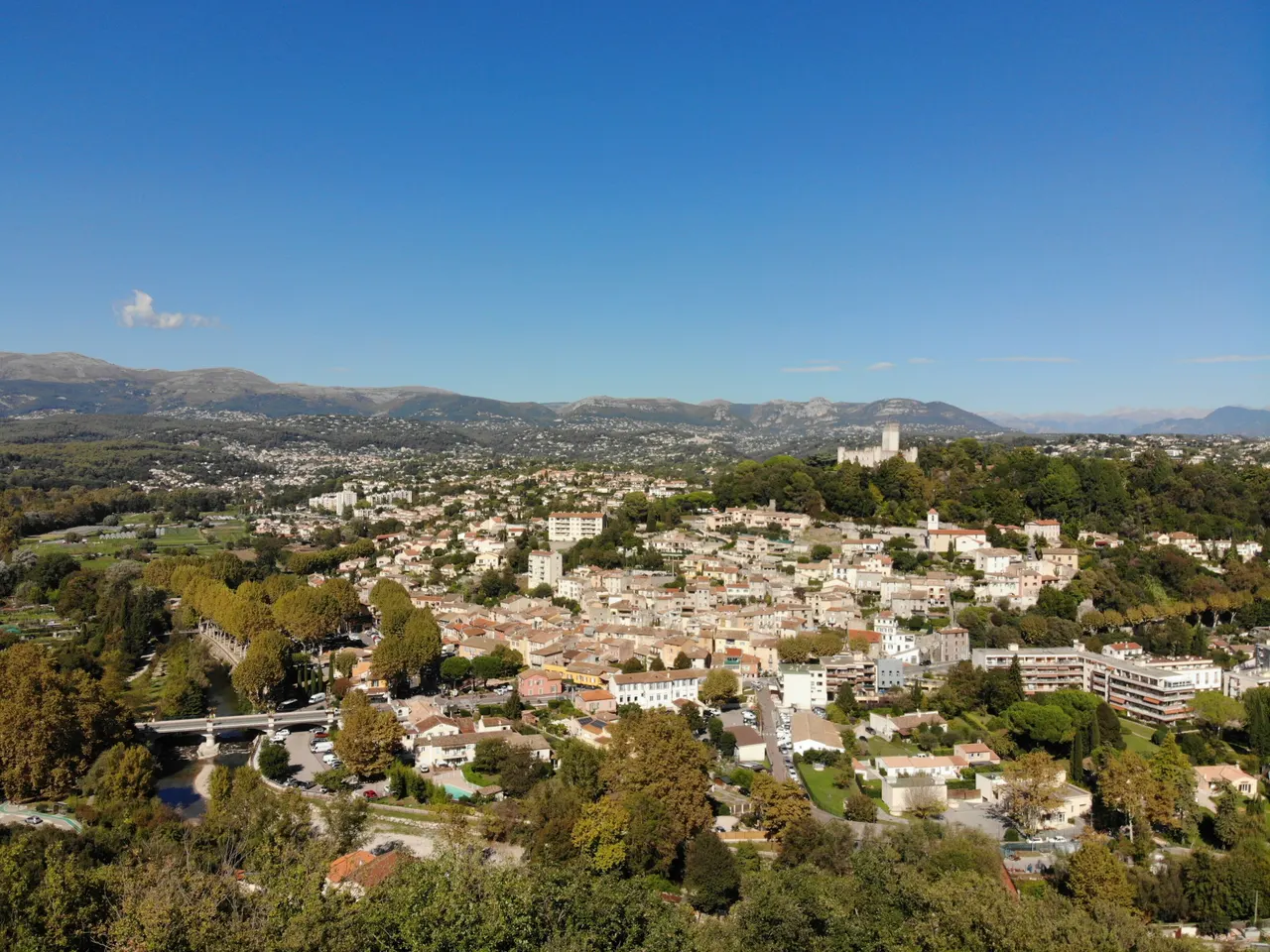Vue aérienne de Villeneuve-Loubet : les toits du bourg le long du Loup et de son pont, le château sur sa colline boisée et les montagnes à l’horizon.