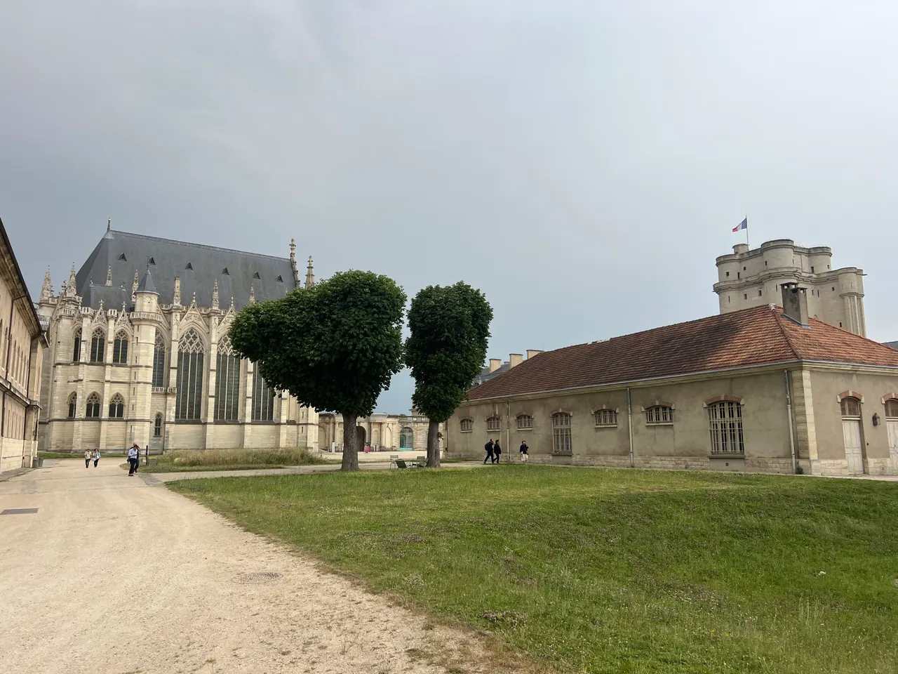 Cour du château de Vincennes : la Sainte-Chapelle gothique à gauche, deux arbres taillés au centre et le donjon au fond à droite, sous un ciel gris